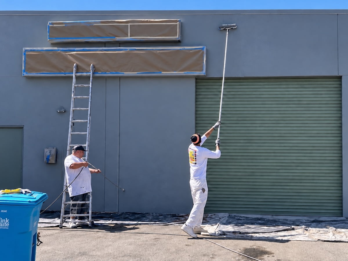Commercial Exterior Spray and Roller Painting Two painters coating a gray commercial building with a sprayer and extension roller, with signs masked above a green roll-up door.